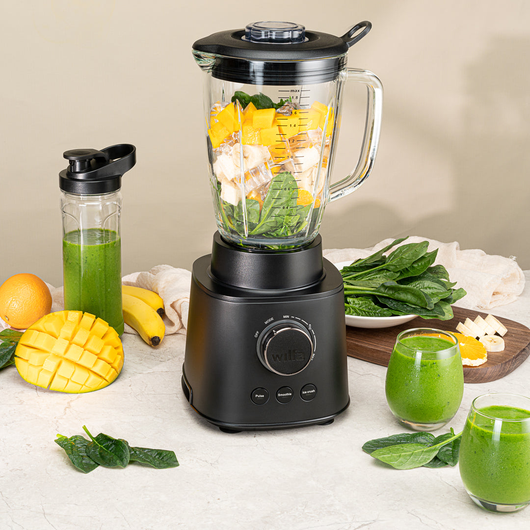Czarny model ESSENTIAL 1800 AUTO z mocnym silnikiem o mocy 1800 W jest wypełniony różnorodnymi składnikami, w tym szpinakiem, bananami i kawałkami mango. Szklany dzbanek z borokrzemianu stabilnie spoczywa na czarnej podstawie wyposażonej w pokrętło regulacyjne i przyciski. Pokrywka jest nałożona, a do niej przymocowane jest czarne narzędzie do mieszania.