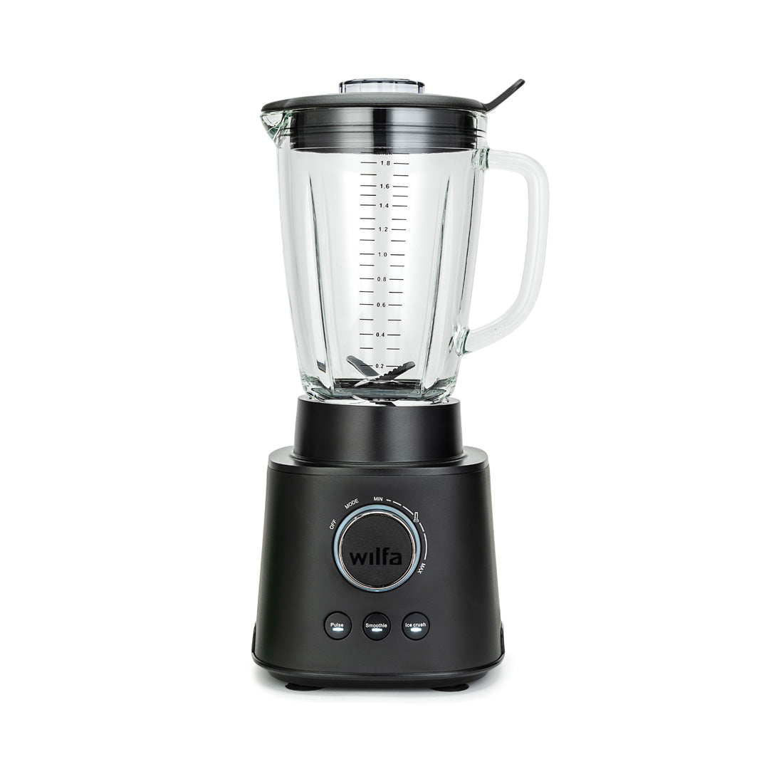 Czarny model ESSENTIAL 1800 AUTO z mocnym silnikiem o mocy 1800 W jest wypełniony różnorodnymi składnikami, w tym szpinakiem, bananami i kawałkami mango. Szklany dzbanek z borokrzemianu stabilnie spoczywa na czarnej podstawie wyposażonej w pokrętło regulacyjne i przyciski. Pokrywka jest nałożona, a do niej przymocowane jest czarne narzędzie do mieszania.