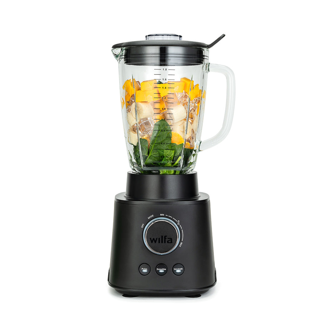 A black ESSENTIAL 1800 AUTO with a powerful 1800 W motor is filled with assorted ingredients, including spinach, bananas, and mango chunks. The borosilicate glass jar sits securely on the black base with a control dial and buttons. The lid is on, complete with a black stirring tool attached to it.