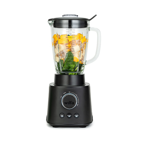 A black ESSENTIAL 1800 AUTO with a powerful 1800 W motor is filled with assorted ingredients, including spinach, bananas, and mango chunks. The borosilicate glass jar sits securely on the black base with a control dial and buttons. The lid is on, complete with a black stirring tool attached to it.