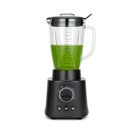 A black ESSENTIAL 1800 AUTO with a powerful 1800 W motor is filled with assorted ingredients, including spinach, bananas, and mango chunks. The borosilicate glass jar sits securely on the black base with a control dial and buttons. The lid is on, complete with a black stirring tool attached to it.