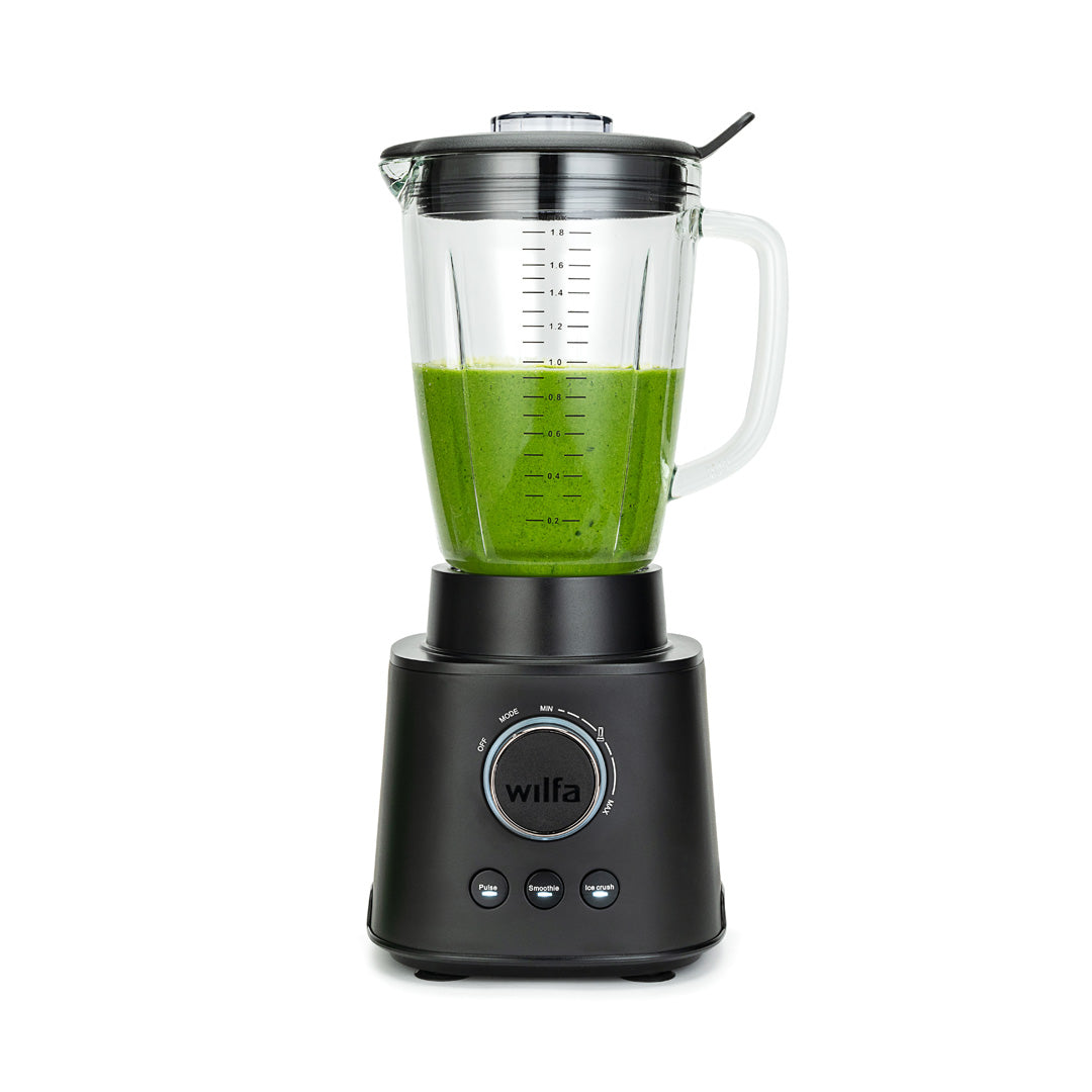 Czarny model ESSENTIAL 1800 AUTO z mocnym silnikiem o mocy 1800 W jest wypełniony różnorodnymi składnikami, w tym szpinakiem, bananami i kawałkami mango. Szklany dzbanek z borokrzemianu stabilnie spoczywa na czarnej podstawie wyposażonej w pokrętło regulacyjne i przyciski. Pokrywka jest nałożona, a do niej przymocowane jest czarne narzędzie do mieszania.