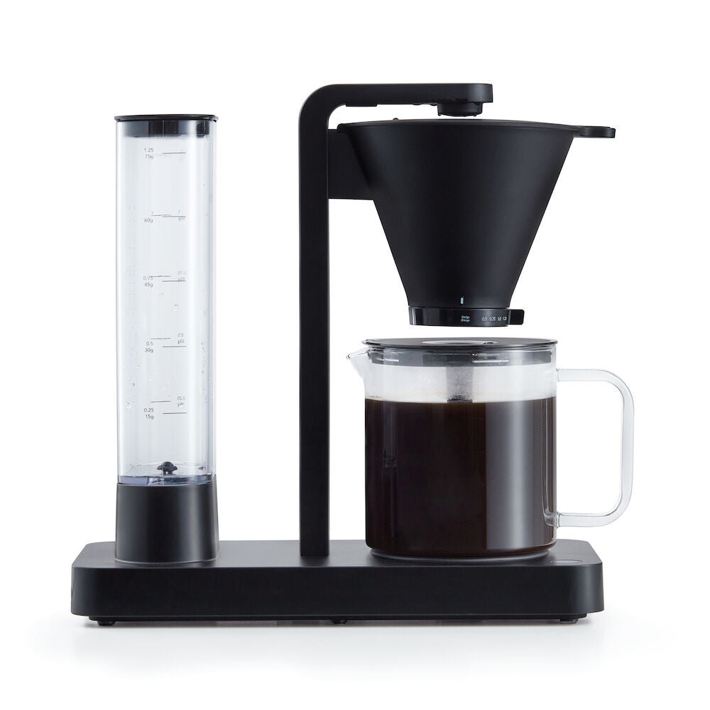 A PERFORMANCE BLACK coffee maker with a transparent water reservoir and a glass carafe filled with coffee. Featuring unique technology for the perfect temperature, it boasts two award badges: one with a laurel wreath indicating "Best i Test Tek.no" and another that reads "Testvinner" with stars and a ribbon symbol.