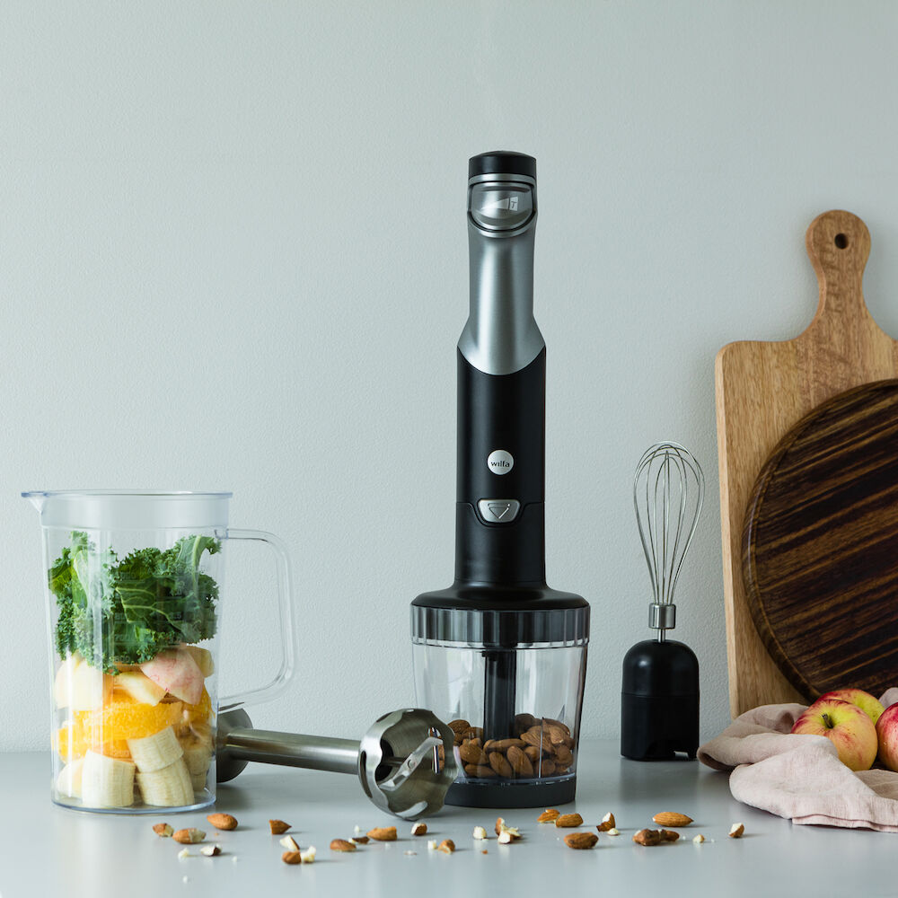 Un juego ESSENTIAL POWER en negro y plateado, con un potente motor y control de velocidad continuo, compuesto por tres elementos: el mango de la batidora, una jarra medidora transparente llena de cebollas rojas picadas y un accesorio transparente para el robot de cocina que contiene verduras picadas, entre ellas cebollas rojas y pimientos verdes.