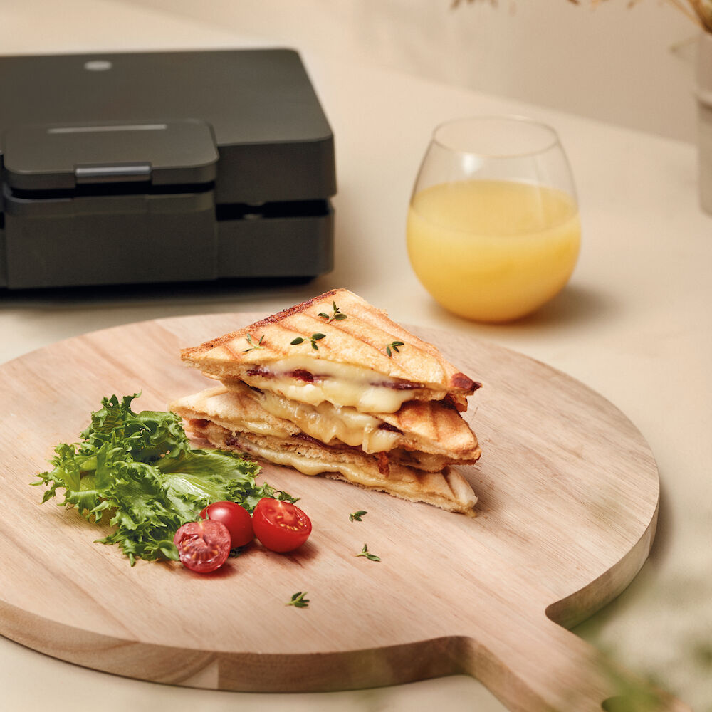 Un appareil à sandwichs ouverts EASY MELT doté de deux compartiments de cuisson triangulaires, avec des surfaces striées et de grandes plaques chauffantes pour griller. L'appareil est noir, doté d'un revêtement antiadhésif, d'une poignée sur le dessus et d'un cordon d'alimentation à l'arrière.