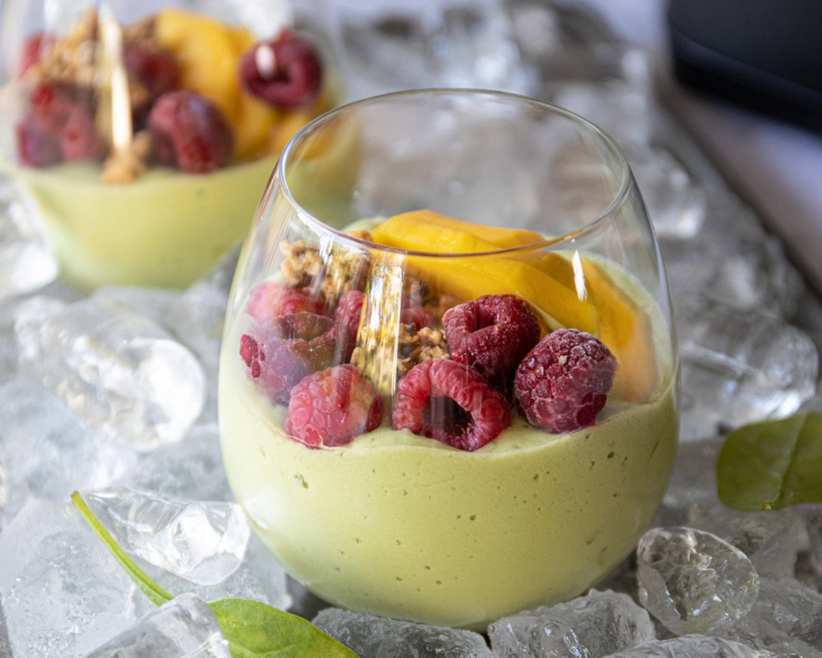 Glass of green smoothie on a tray of ice cubes, topped with raspberries, granola and mango