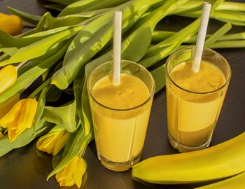 Two glasses of mango banana smoothie with straws, yellow tulips and banana