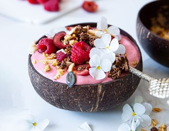Pink raspberry and banana smoothie bowl in a coconut shell topped with raspberries, granola and flowers