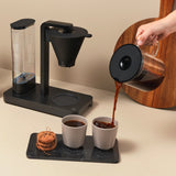 A PERFORMANCE BLACK coffee maker with a transparent water reservoir and a glass carafe filled with coffee. Featuring unique technology for the perfect temperature, it boasts two award badges: one with a laurel wreath indicating "Best i Test Tek.no" and another that reads "Testvinner" with stars and a ribbon symbol.