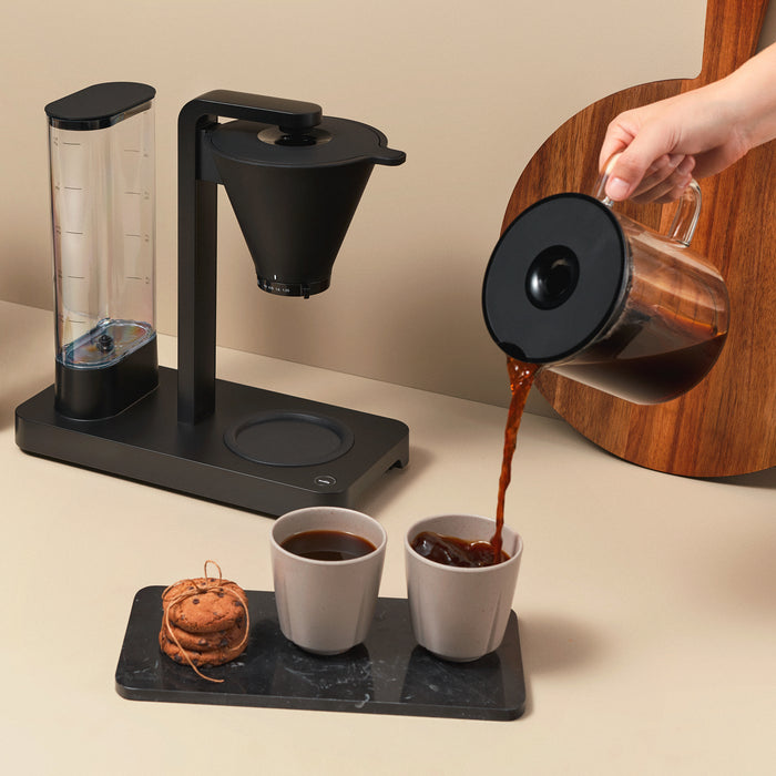 A PERFORMANCE BLACK coffee maker with a transparent water reservoir and a glass carafe filled with coffee. Featuring unique technology for the perfect temperature, it boasts two award badges: one with a laurel wreath indicating "Best i Test Tek.no" and another that reads "Testvinner" with stars and a ribbon symbol.
