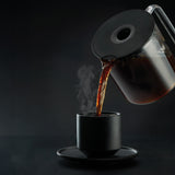 A PERFORMANCE BLACK coffee maker with a transparent water reservoir and a glass carafe filled with coffee. Featuring unique technology for the perfect temperature, it boasts two award badges: one with a laurel wreath indicating "Best i Test Tek.no" and another that reads "Testvinner" with stars and a ribbon symbol.