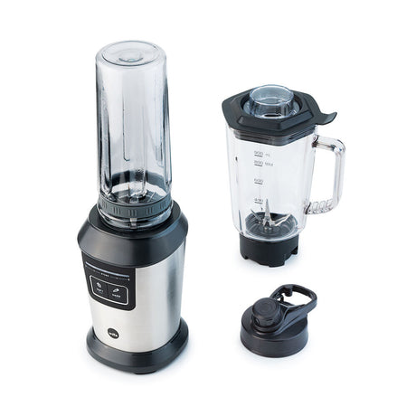 A stainless steel sports blender with a transparent pitcher filled with blueberries, spinach, and other fruits. Next to it is an ACTIVLIFE STEEL 2go bottle containing a dark purple smoothie topped with a secure lid. A small bunch of blueberries and a couple of mint leaves are placed nearby.