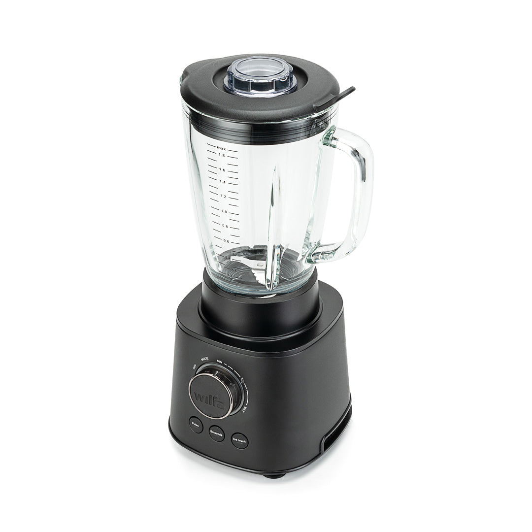 A black ESSENTIAL 1800 AUTO with a powerful 1800 W motor is filled with assorted ingredients, including spinach, bananas, and mango chunks. The borosilicate glass jar sits securely on the black base with a control dial and buttons. The lid is on, complete with a black stirring tool attached to it.