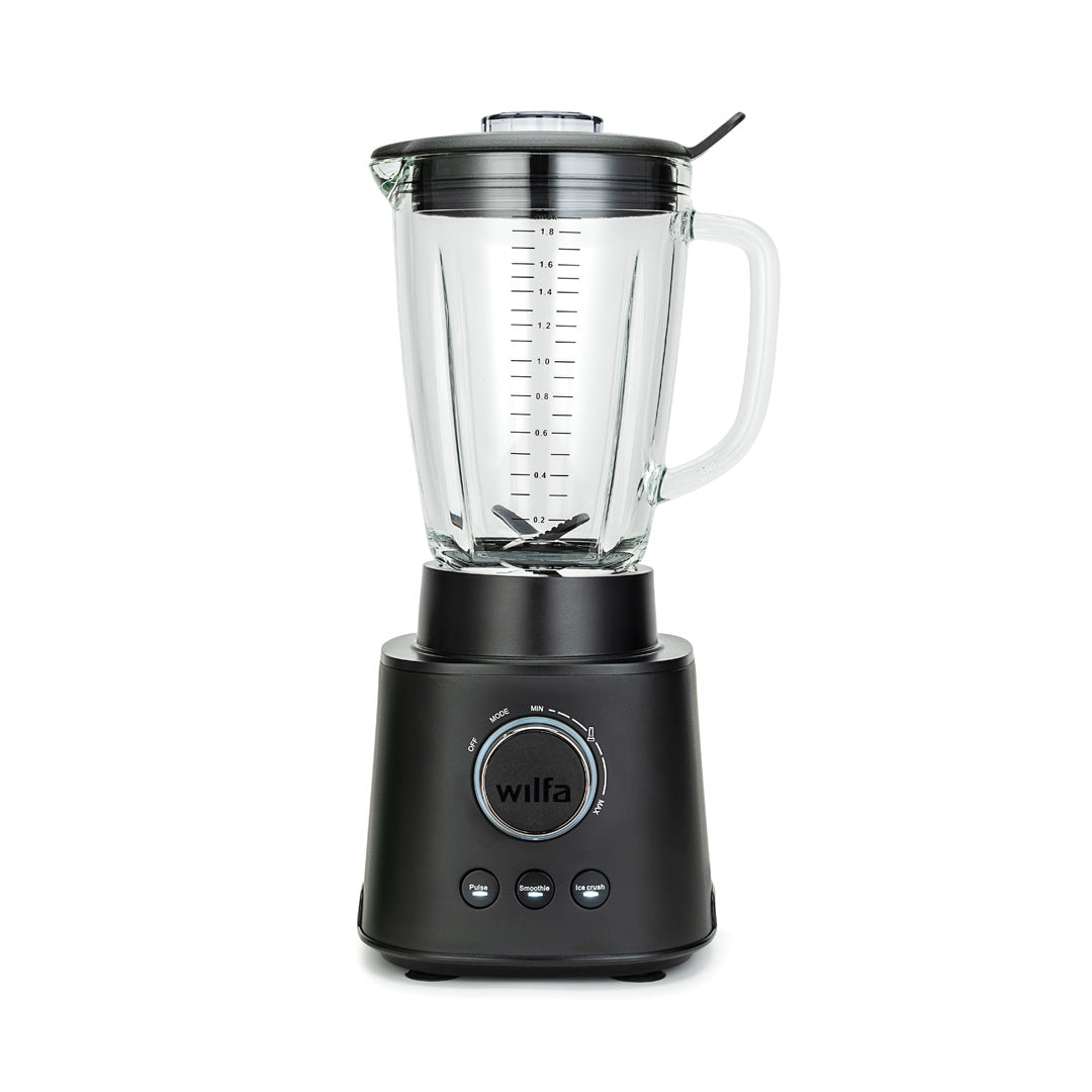 A black ESSENTIAL 1800 AUTO with a powerful 1800 W motor is filled with assorted ingredients, including spinach, bananas, and mango chunks. The borosilicate glass jar sits securely on the black base with a control dial and buttons. The lid is on, complete with a black stirring tool attached to it.