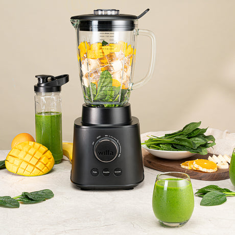 A black ESSENTIAL 1800 AUTO with a powerful 1800 W motor is filled with assorted ingredients, including spinach, bananas, and mango chunks. The borosilicate glass jar sits securely on the black base with a control dial and buttons. The lid is on, complete with a black stirring tool attached to it.