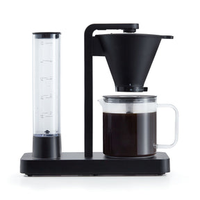 A PERFORMANCE BLACK coffee maker with a transparent water reservoir and a glass carafe filled with coffee. Featuring unique technology for the perfect temperature, it boasts two award badges: one with a laurel wreath indicating "Best i Test Tek.no" and another that reads "Testvinner" with stars and a ribbon symbol.