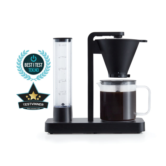 A PERFORMANCE BLACK coffee maker with a transparent water reservoir and a glass carafe filled with coffee. Featuring unique technology for the perfect temperature, it boasts two award badges: one with a laurel wreath indicating "Best i Test Tek.no" and another that reads "Testvinner" with stars and a ribbon symbol.