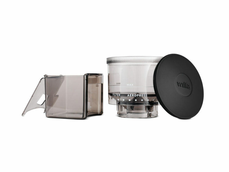 Image of an AeroPress coffee maker with its components separated. From left to right: the gray plunger, the transparent brew chamber with white measurement markings and a silver top, and the black filter cap. The components are arranged on a plain white background, next to a sleek SVART BEAN AND COFFEE CUP W/LID.